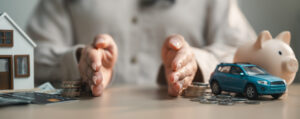 woman's hands separating figurines: a house, car, and piggy bank, and cash