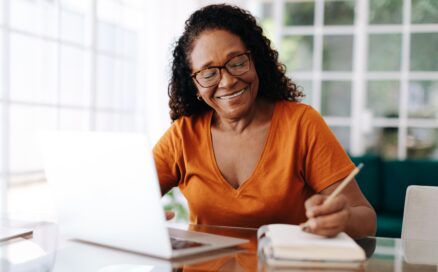 Happy senior woman writing in a notebook with laptop in front of her